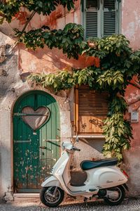 A white Vespa scooter parked by a charming old door with a heart design, surrounded by lush greenery, evoking a quaint Italian vibe.