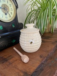 This beautiful ceramic honey pot features horizontal ridges to create a beehive effect, a hand painted bee design and a simple lid with a stopper. Use its traditional dibber to effortlessly drizzle honey over all your favourite foods.  It has been painted using honey toned speckle underglaze paints and then glazed and fired in my studio kiln.  Dimensions: Pot & Lid:  11cm (H) x 9cm (W) x 9cm (D) Dibber: 11.5cm (H) x 2cm (D) Thank you for taking the time to look at my range. It really does mean a