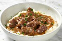 Angled view of beef tips on mashed potatoes, topped with herbs in a white and brown speckled bowl.