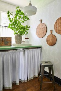 Decorative cheese boards hang from a light gray wallpapered wall to the side of a green countertop accented with pleated curtains hung in front of shelves, while a Tolix stool sits in front of it facing a window covered in white cafe curtains.