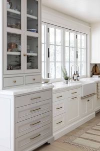 In our white and grey kitchen design, we added a built-in dining hutch painted in a greige accent color to the kitchen cabinetry. Our clients have lots of heirloom antique glassware that they use for entertaining and special occasions. The glass cabinet doors allow for them to be on prime display. Brass cabinet hardware and honed marble countertops complete the kitchen cabinetry design!