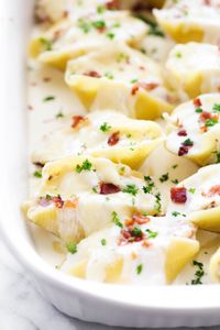 Large pasta shells stuffed with a cream cheese mixture, drizzled with a white cream sauce and garnished with bacon crumbs and parsley in a white casserole pan.