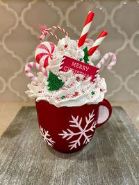 Handcrafted with care, this approximate 5-inch tall by 3-inch wide festive Christmas ornament features a red mug adorned with glittering snowflakes, overflowing with fluffy faux whipped cream, shimmering red and white candy canes, and topped with a "Merry Christmas" sign. Peppermint candies and tiny Christmas trees add a whimsical, playful vibe, while sparkling glitter gives it that extra holiday shine. This ornament is the perfect addition to any Christmas tree, tiered tray, or kitchen decor, making it an eye-catching statement piece. It's also a charming gift for anyone who loves holiday decor or baking-inspired decorations. Whether you're decorating for a cozy night in or hosting a festive gathering, this whimsical fake bake will bring warmth and cheer to your Christmas setup!