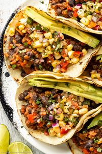 Overhead, close-up photo of black bean tacos topped with corn salsa and slices of avocado on a white plate with fresh lime wedges next to the plate.