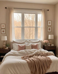 This soft and cozy bedroom features a plush bed layered with white and blush pillows, along with a light throw blanket. The large windows fill the room with natural light, while the neutral walls and minimal decor create a serene and relaxing atmosphere. Wooden bedside tables with lamps and floral accents add warmth and a touch of elegance. #CozyBedroom #BlushAccents #BedroomDecor #NeutralInteriors #SereneSpace