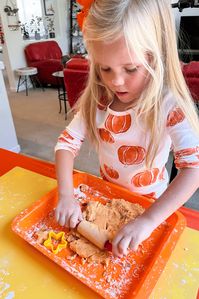 A young girl uses a small rolling pin to flatten the 2-ingredient pumpkin dough on an orange tray, getting creative during a preschool sensory activity.