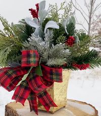 Winter centerpiece with a plaid bow, gold container, and evergreens. Put this out all winter long! #pinklimewreaths #christmastabletopcenterpiece #winterarrangements #christmasdecor #christmastabledecoration #holidaycenterpiece #christmasflowerarrangement #fauxchristmasarrangements