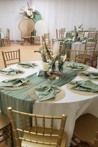 Embrace rustic charm at your event tables. White tablecloths and sage green table runners create a serene base with gold woven placemats and stylishly folded sage green napkins on top. The center hosts candles in themed holders, surrounded by rustic flower arrangements. A Chiara stand backdrop, draped in white and sage green spandex, showcases a boho-themed flower arrangement, completing the rustic allure for an enchanting celebration. 🌿✨ #RusticStyle #EventDecor