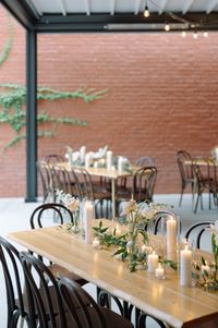 The modern ambiance flipped from the ceremony to a more casual setting for their grazing style dinner. Simple tablescapes consist of a mix of pillar and votive candles, dark greens and bud vases with white blooms. Exposed wood tables with dark mahagony bentwood chairs.   Sera Petras Photography. Days and Eves Wedding Planning. CommonHouse RVA Catering and Venue. Fleurish and Bloom fresh buds and Candles.