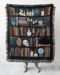 "Introducing a charming and eerie cotton throw blanket that will add a touch of magic to your home decor! Designed to resemble a witch's bookshelf, this blanket features a captivating mix of skulls, potions, books, a crystal ball, and a snoozing black cat. Perfect for Halloween or as a year-round accent for your favorite dark cottagecore witch, this woven blanket is crafted with colored threads for long-lasting durability. Snuggle up with this whimsical and spooky throw and let the enchanting at