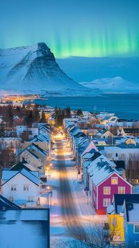 A photo of Reykjavik, Iceland during the blue hour, with snow-covered streets and colorful houses illuminated by soft yellow streetlights. The city is quiet, with the backdrop of snow-covered mountains and the icy waters of the Atlantic Ocean. The sky glows faintly with the Northern Lights in the distance, creating a magical and serene atmosphere.