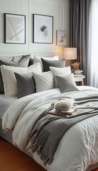 The image depicts a cozy, well-decorated bedroom. The bed is made up with a white comforter and several decorative pillows in shades of grey and white, featuring various textures and patterns. A grey throw is draped artfully over the bed. On the bed is a stack of books with a small bowl and a soft object on top. Beside the bed is a nightstand with books, a lamp, and dried flowers in a vase. The wall is decorated with framed abstract art, and grey curtains hang by the window, allowing soft natural light to filter into the room. #BedroomDecor #CozyBedroom #NeutralColors #InteriorDesign #HomeDecor #CalmingSpaces #ModernBedroom #GreyAndWhiteDecor #ComfortableLiving #BedroomInspiration #homebedroomrefresh #bedroomideas #cozybedroom #chillroom #apartmentbedroom #bedroomaesthetic