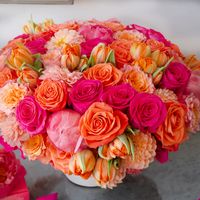 Indulge in a vibrant and meticulously crafted floral arrangement that will enchant your senses. It features a lush gathering of the vivid and bold "Orange Crush" roses, the charming "Free Spirit" roses, complemented by the gentle beauty of peach dahlias. To further enhance this masterpiece, there are delicate tulips and striking hot pink peonies, adding a sense of joy and celebration. This lively ensemble is a true "Happy Fiesta" of flowers, artfully designed in a pave style. It stands at an imp