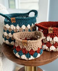 Three crocheted Granny Stitch Baskets in varying sizes and colors sit on a wooden surface. Each basket has a label reading "Mo's" and showcases a mix of teal, beige, and rust hues.