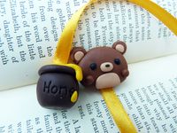 Isn't it funny how bears like honey? This beautiful bookmark will brighten up anyone's day!  A handmade polymer clay bear sits on top of your book, proudly marking your page, while a colourful ribbon runs down to a little honey pot, that's overflowing with sweetness!!   Make a perfect gift for anyone who loves books, or a perfect indulgence for a bookaholic!   Each bookmark will be delivered in its own organza bag!  Please note: each bookmark is handmade, so may vary slightly from the picture, but that just makes them all the more unique! Why not take home the Bear and Honey Pot bookmark today and start an adventure together!