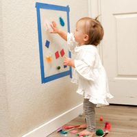 A toddler sticks shapes to a sheet of contact paper attached to a wall with the sticky side out