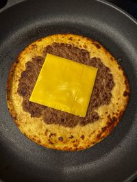 Slice of American cheese melting in the center of a freshly smashed burger on a golden brown tortilla shell in a black non-stick skillet.