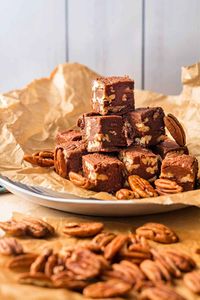 A plate with chocolate fudge with pecans in it all stacked up. There are loose pecans on beige parchment in the foreground.