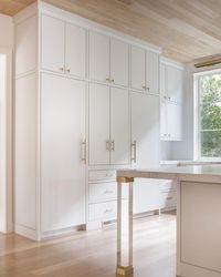 Brass and lucite legs, placed on gray wash wood floors under an oak plank ceiling, accent a white island topped with a gray and white marble countertop facing white concealed fridges placed both under and on either side of white flat front cabinets adorning Lewis Dolan Bar Pulls and Knobs.