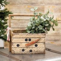 The Lath Snowman Tote is a rustic wooden tote decorated with distressed cream paint. The tote is rectangular in shape, featuring a sturdy wooden handle and rough, worn edges. The tote displays black painted details and an orange carrot nose, creating a smiling snowman face. The side of the tote includes a decorative re