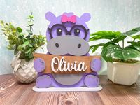 This adorable little hippo is ready to be personalized with any name. Choose between options with or without a bow and select your choice for the color of the bow.  This personlized name stand sit easily on any surface including: desks, shelves, night stands, dressers, and more. Our adorable hippo measures approx 10.5" tall and is made out of wood. Each hippo is hand painted with acrylic paint and best for indoor use.  Please let us know if you have any questions or special requests and we will