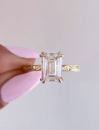 Esmeralda is an elevated, vintage inspired solitaire that captures the essence of timeless romance. This piece features a breathtaking emerald cut diamond hugged by a hidden halo and double prongs. The band showcases intricate hand-engraved regal details, reminiscent of vintage artistry, ensuring that each glance reveals a new layer of beauty.