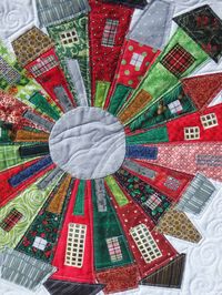 "\"Christmas Village\" is an original quilt using the traditional Dresden plate pattern. The segments have been used to create a Christmas village. Done in shades of red and green, this 21 by 22 wall hanging is unique in the little details such as the windows and doors. The houses are outline quilted and there are spirals quilted in the sky. The back of the quilt features a Christmas print and the necessary corner flaps for hanging with a dowel rod. The green double fold binding is hand stitched