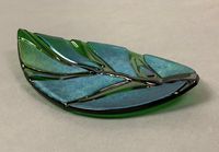 "This kiln-formed leaf tray combines green glass with panels of metallic iridescent glass. The gently sloping leaf tray is perfect for display, catching your keys, or for serving up crackers at your next party. Handmade fused glass tray is kiln fired, fire polished, and properly annealed for strength and durability. Hand washing recommended. Do not use tray in microwave. Tray is available in 2 sizes: Large: 12\" x 7\" Small: 6\" x 4\" As each tray is individually handmade, actual tray may vary s