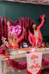 bride in pink sequin jacket and shorts with bride in matching pink sequin dress sit at sweetheart table during wedding reception at Fig House Los Angeles | Feathered Arrow Studio #losangelesweddingplanner #lgbtqwedding #colorfulweddings