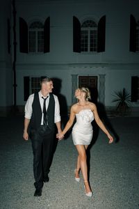  Looking for the perfect after-party dress inspiration? This bride nailed it with a stunning, short white dress featuring a bold floral detail. Paired with stylish heels, she exudes effortless elegance and fun. The groom complements her look with a relaxed yet sharp black vest and tie ensemble.