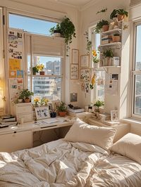 A cozy dorm room with a bed, desk, and plenty of plants. The room is decorated with artwork and has a large window that lets in plenty of natural light. The bed is neatly made and has a white comforter. The desk is cluttered with books and other items, giving the room a lived-in feel. The room is filled with various potted plants, adding a touch of greenery to the space.