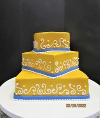 Three tier square cake in gold buttercream with white scrollwork and blue fondant bands