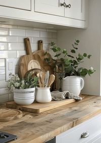 Elevate your farmhouse kitchen with this stylish counter setup. Enjoy a blend of plant decor, a wooden cabinet, and white walls for a cozy, charming look.