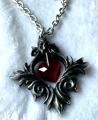 "An 1 ½\" long antiqued pewter pendant of a baroque scroll frame surmounted by a small skull and with a red Austrian crystal heart suspended at its centre. The pendant comes with a velvet gift pouch. Made by Kiss of a red rose with help from Alchemy England 1977."