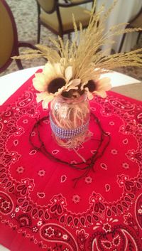 Country themed party table decor for circular tables. Bandana topped with centerpiece made with fake sunflowers and wheat from Michaels. Filled with straw-like gift filler. Mason jars wrapped with gingham, juke string tied into a bow. Barbed wire string from Oriental Trading. #country #western #masonjar #gingham #bandana #paisley #sunflowers #wheat #centerpiece #tabledecor #party #oklahoma #barbedwire #makeitwithmichaels