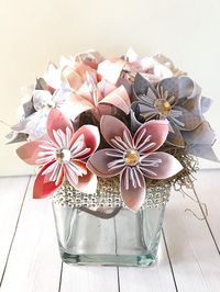 A clear square glass vase measuring 3.9”x3.9” is topped with origami kusudama and lily flowers. Each one is made to order with your custom colors and choice of paper. The top edge of the glass is finished with your choice of rhinestones or ribbon. Inside the vase is a branch with 2