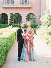 Bridesman in blush bowtie and black tux with Bride wearing a Marchesa blue and pink floral long sleeve couture gown with floral appliques. Large bridal bouquet of mostly pink tones roses, peonies, ranunculus, and sweet peas with long bright pink and blush and mauve silk ribbons. Fairmont Grand del Mar wedding published on Martha Stewart Weddings