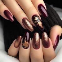 The nails are painted in a deep burgundy shade. Each nail features delicate golden maple leaves, some outlined in black for added depth. The tips of the nails have a subtle gold glitter gradient, adding a shimmering effect that catches the light beautifully.