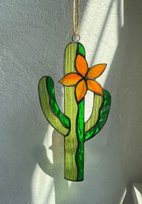 This Stained Glass Desert Cactus with Flower Suncatcher brings the beauty of the desert into your home. The textured cathedral (translucent) green glass resembles the look of a cactus and the deep orange flower petals create a warm glow when the sun shines through☀️🌵 We recommend not using suction cups due to them coming loose over time. Hang from a nail or window lock.