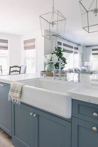 A Shaw farm sink paired with a chrome hook and spout faucet is fixed to blue island painted Benjamin Moore Britannia Blue are accented with chrome knobs and a white quartz countertop lit by two Darlana Lanterns.