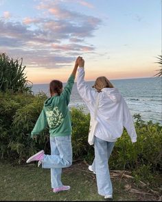 two people standing on top of a lush green field next to the ocean with their arms in the air