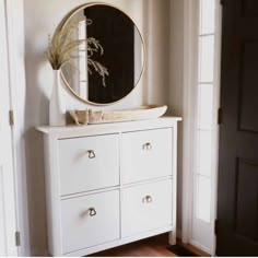 a white dresser with a round mirror above it and a plant in a vase on top