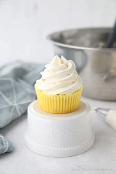 a cupcake with white frosting sitting on top of a cake plate next to a mixing bowl