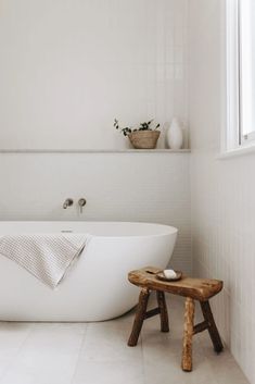 a white bath tub sitting next to a wooden stool in a bathroom under a window