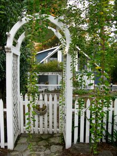 a white picket fence with an arch in the middle and green plants growing on it