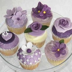 cupcakes decorated with purple and white frosting on a glass platter topped with flowers