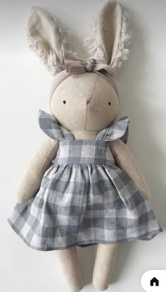 a stuffed rabbit wearing a blue dress and bow on it's head, sitting against a white wall