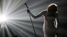 a woman standing in front of a microphone on stage with bright lights behind her,