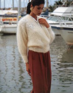 a woman in a white sweater and brown skirt standing on the dock next to boats