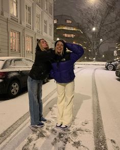 two women are standing in the snow on a street at night with their arms around each other